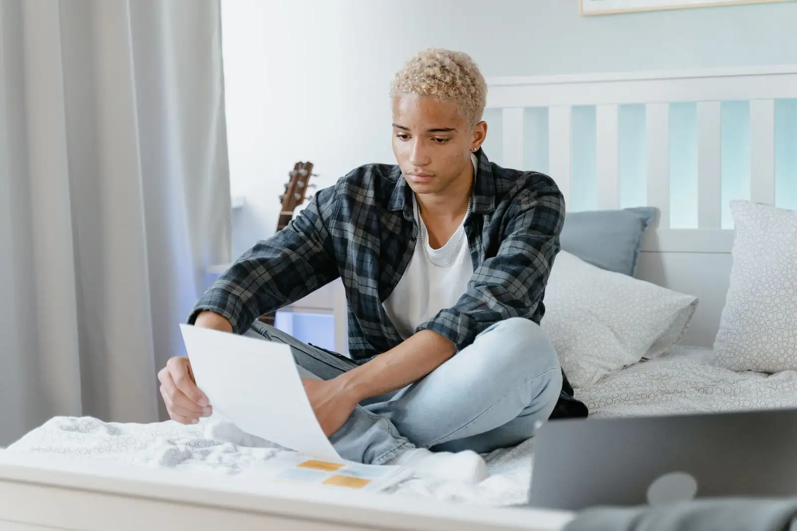 Jeune personne installée sur le lit de sa chambre, un document à la main, à côté d'un ordinateur portable.