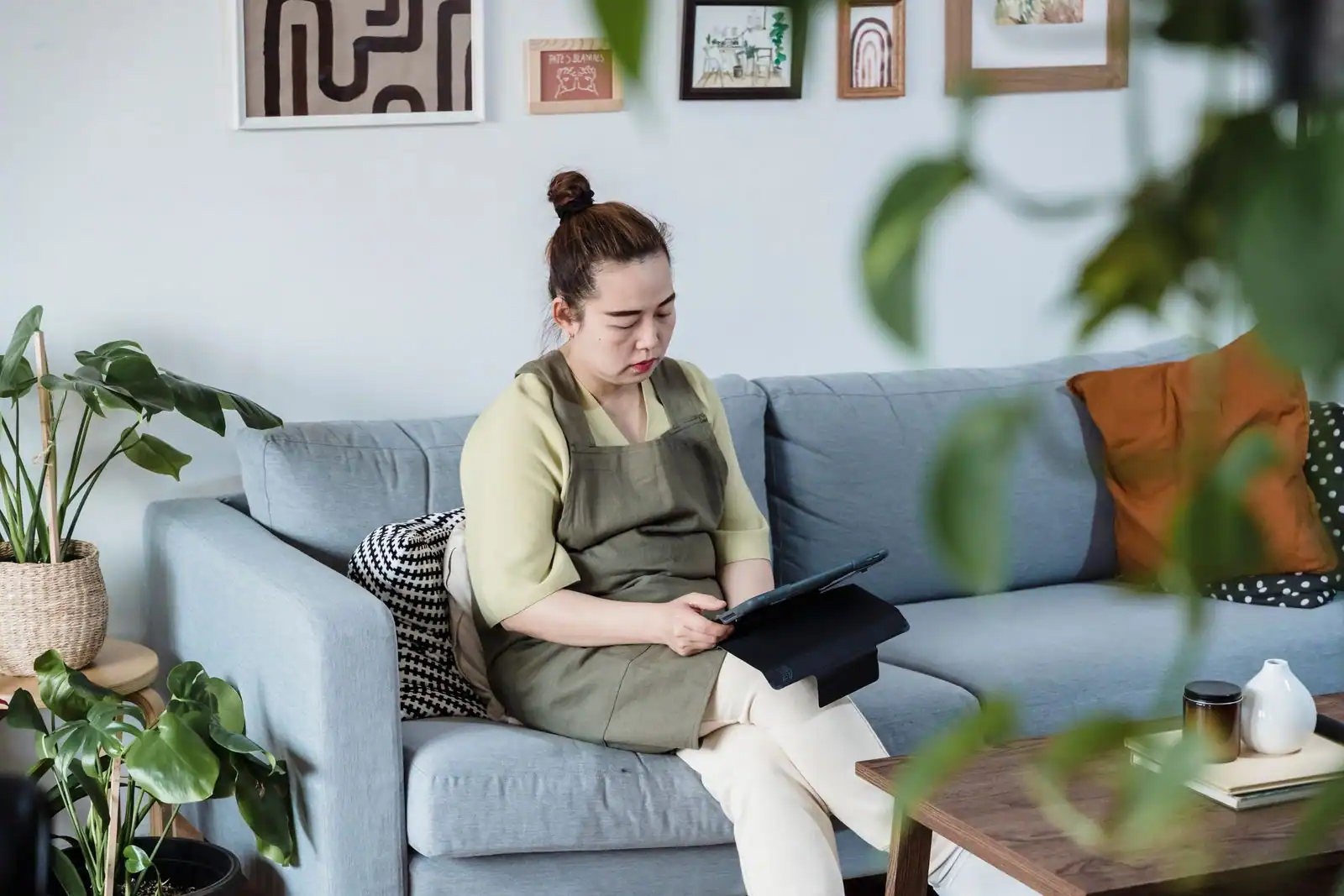Personne utilisant une tablette, assise sur le canapé de son salon décoré de cadres et de plantes.