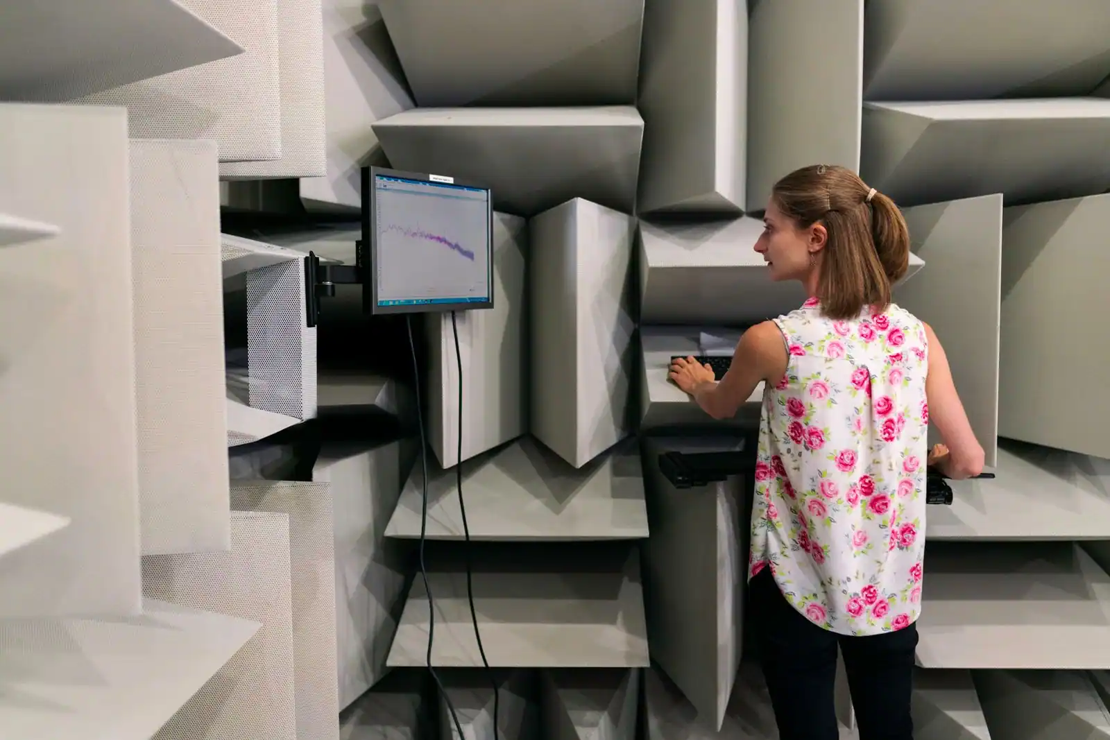 Intérieur d'une chambre anéchoïque tapissée de dièdres absorbants, avec une technicienne devant un écran de mesure
