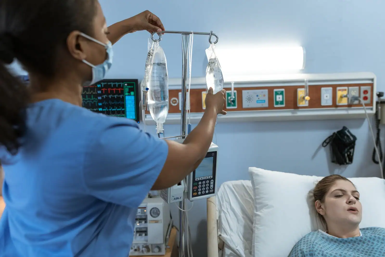 Soignante installant des poches de perfusion intraveineuse près d'un patient, dans une chambre d'hôpital.