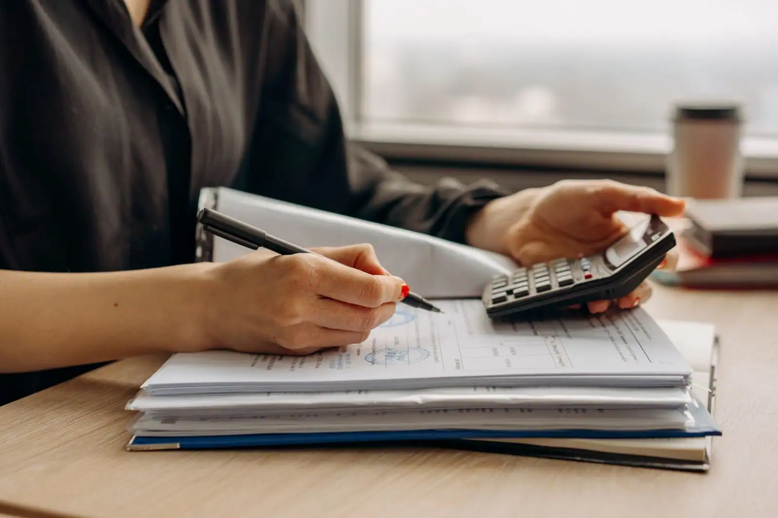 Personne calculant des chiffres avec une calculatrice et un stylo sur une pile de documents posée sur un bureau.