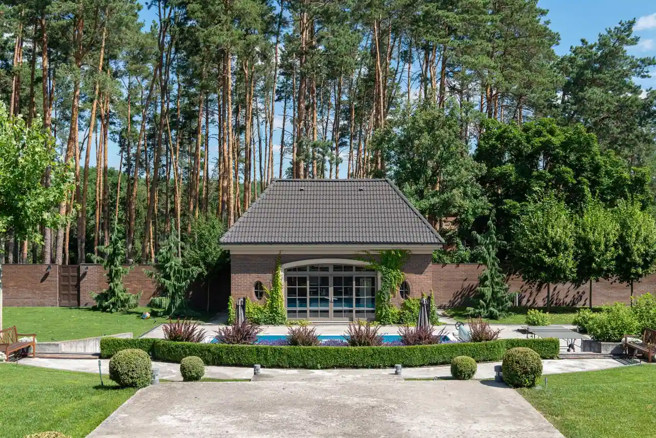 Une piscine bleue intégrée dans un jardin paysager taillé, avec un pool house et des pins en arrière-plan.