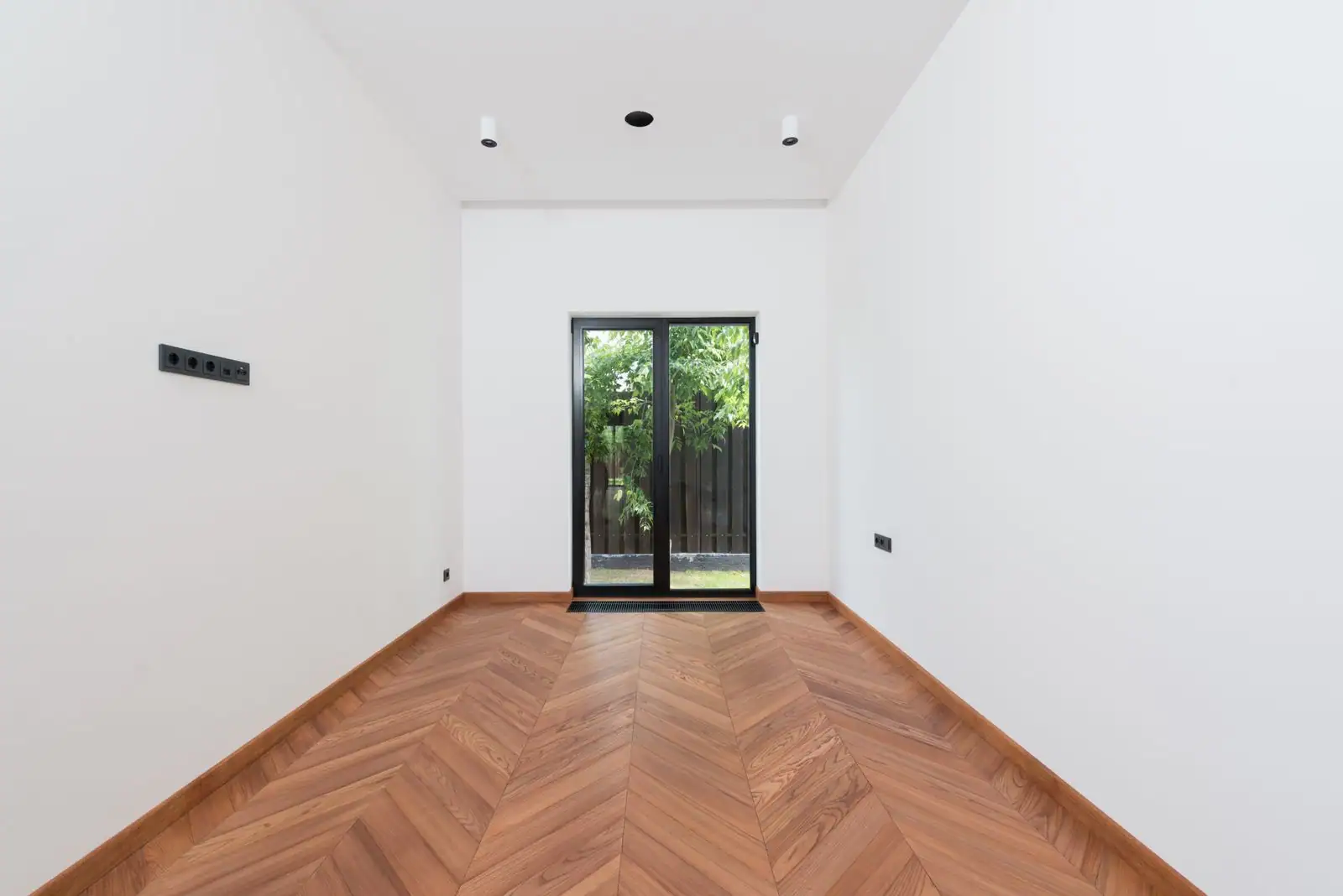Pièce vide fraîchement rénovée avec parquet en chevron, murs blancs et porte vitrée ouvrant sur un jardin