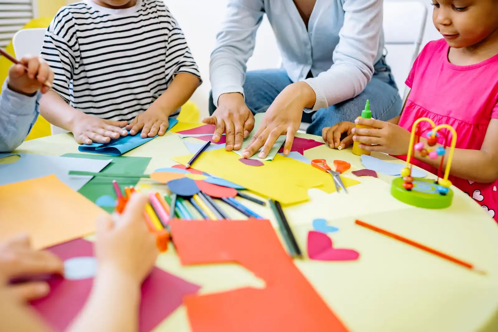 Des enfants assemblent des papiers de couleur autour d'une table, les mains d'un adulte posées à côté