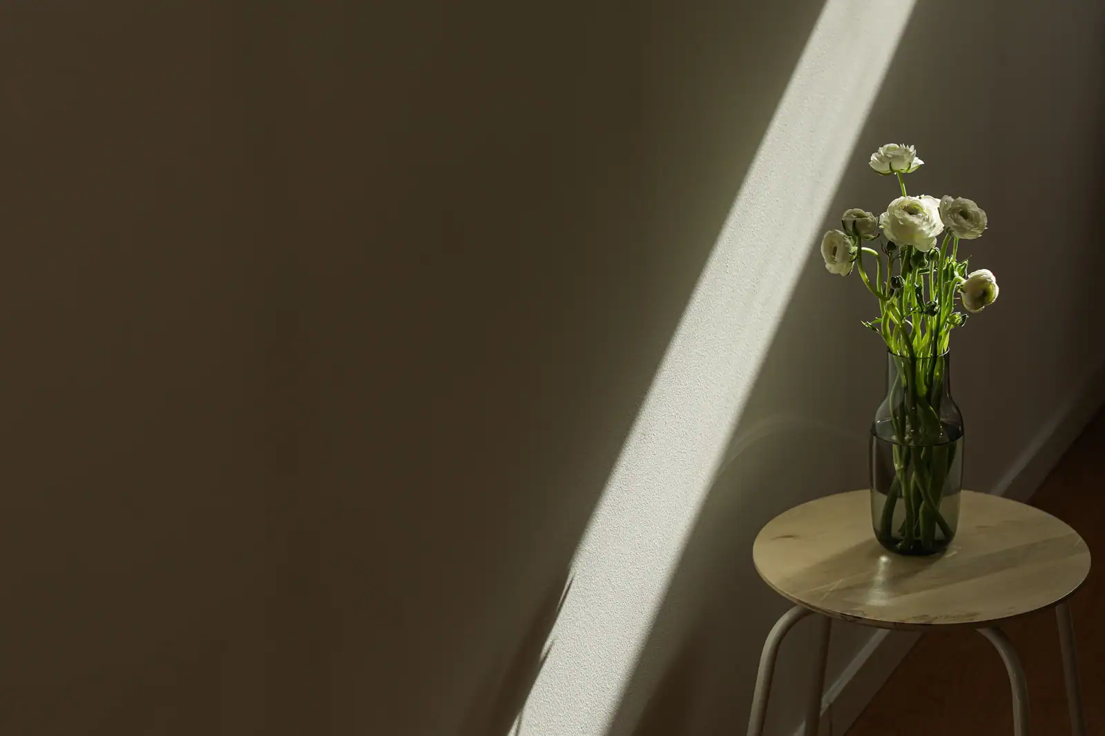 Petit bouquet de fleurs blanches dans un vase sombre, posé sur un tabouret en bois dans une pièce nue.