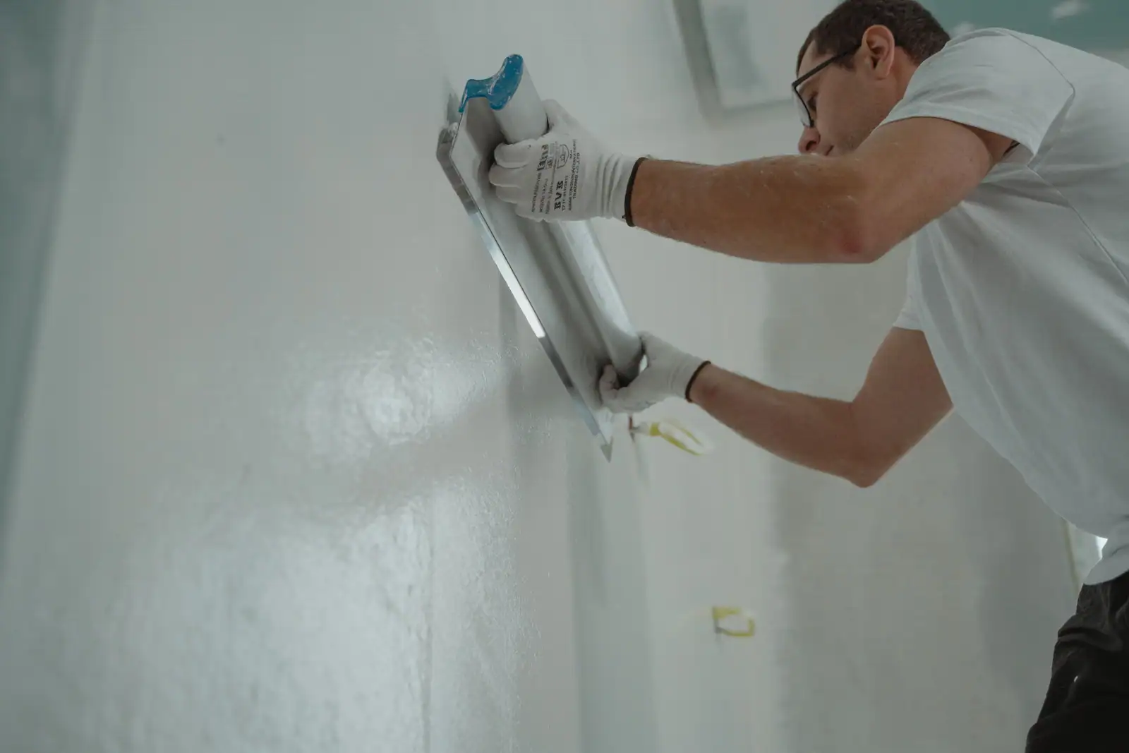 Un professionnel lisse un enduit sur un mur intérieur avec un large platoir, gants et lunettes de protection