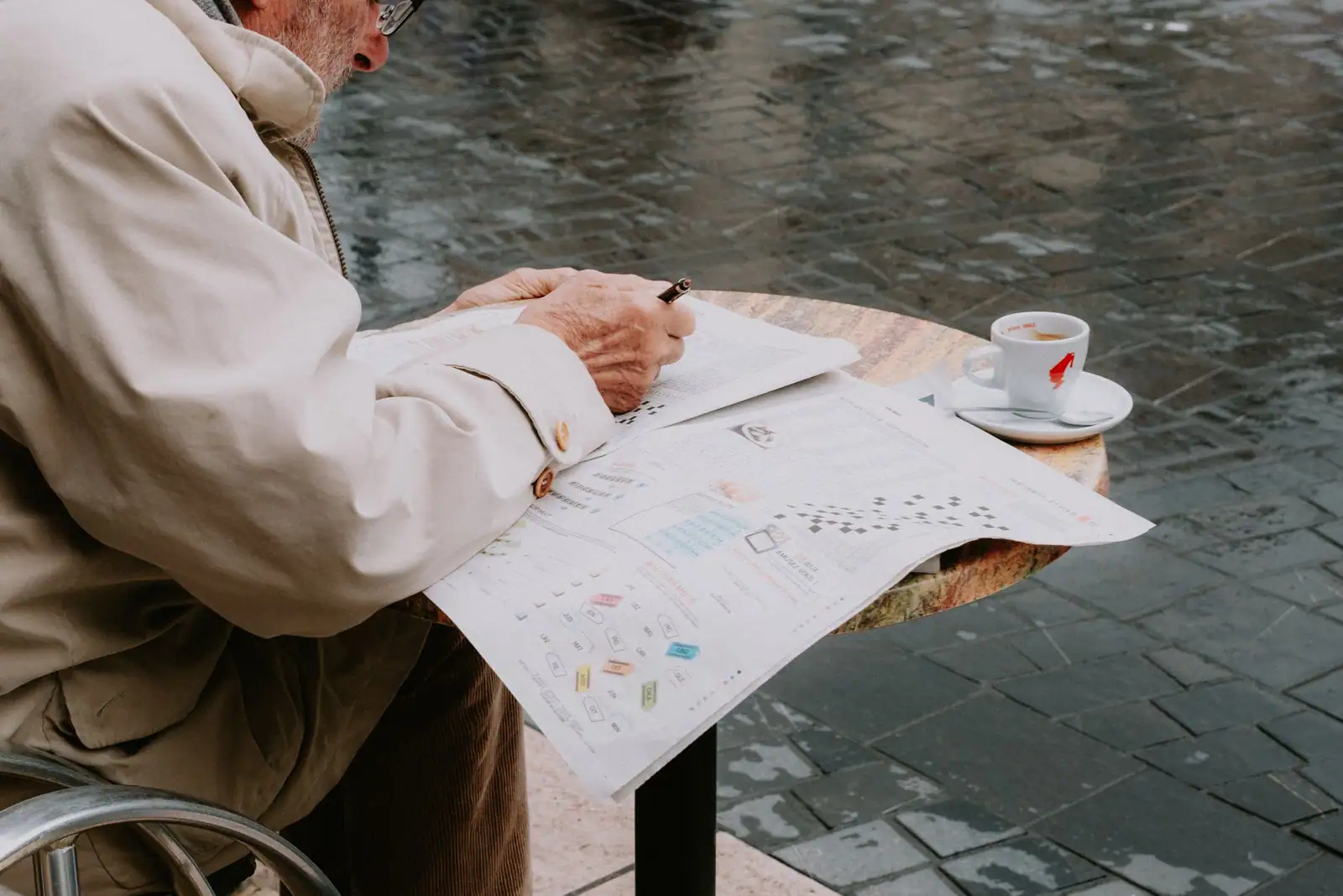 Homme attablé à la terrasse d’un café, stylo en main, penché sur la page jeux d’un journal déplié