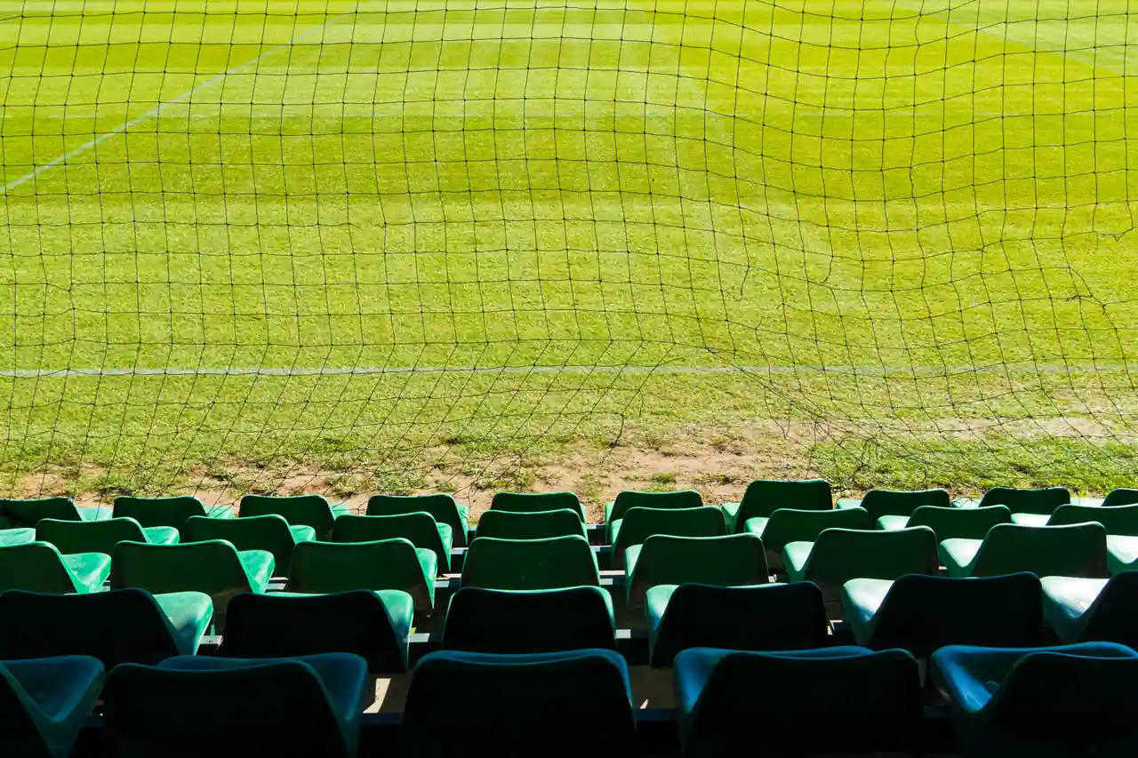 Rangées de sièges vides derrière un filet de protection, face à la pelouse d'un stade de football