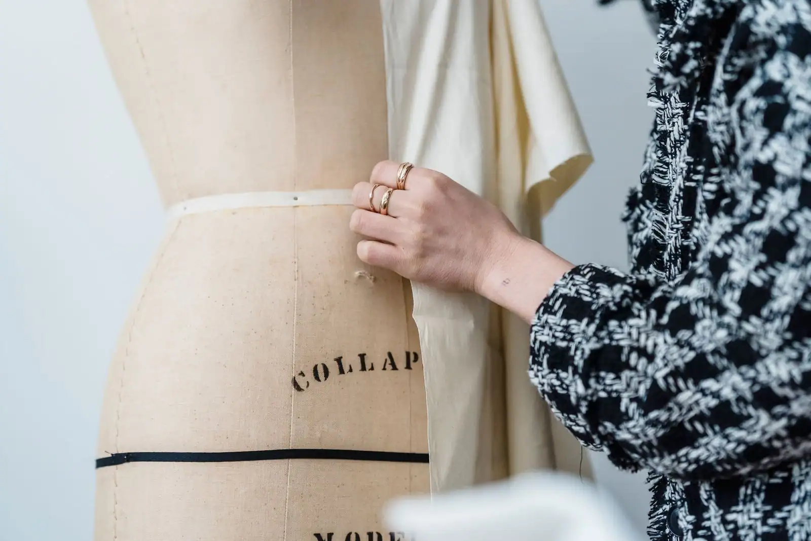 Une main épingle un tissu écru sur un mannequin de couture toilé, marqué de lignes de repère noires