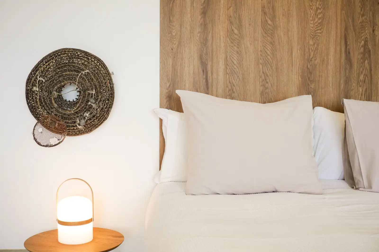Tête de lit en bois clair, literie écrue et pièce en fibres tressées au mur, près d’une lampe ronde
