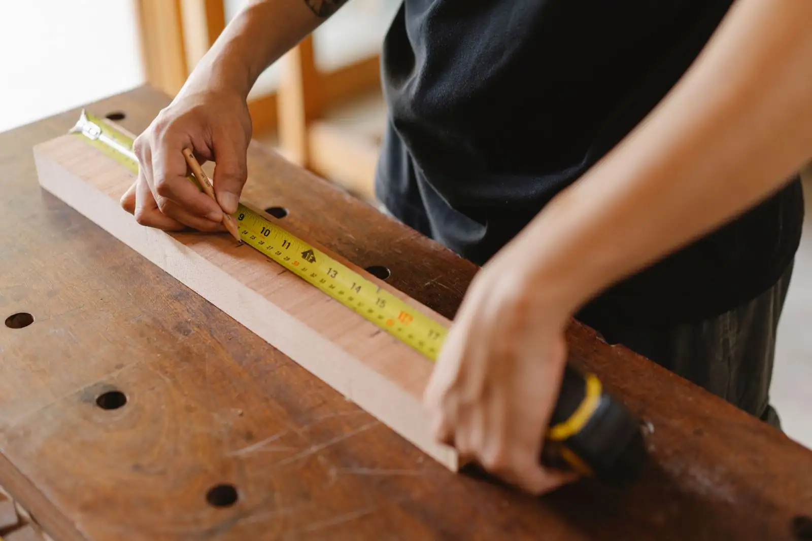 Menuisier mesurant une planche au mètre ruban sur un établi, un crayon de traçage entre les doigts