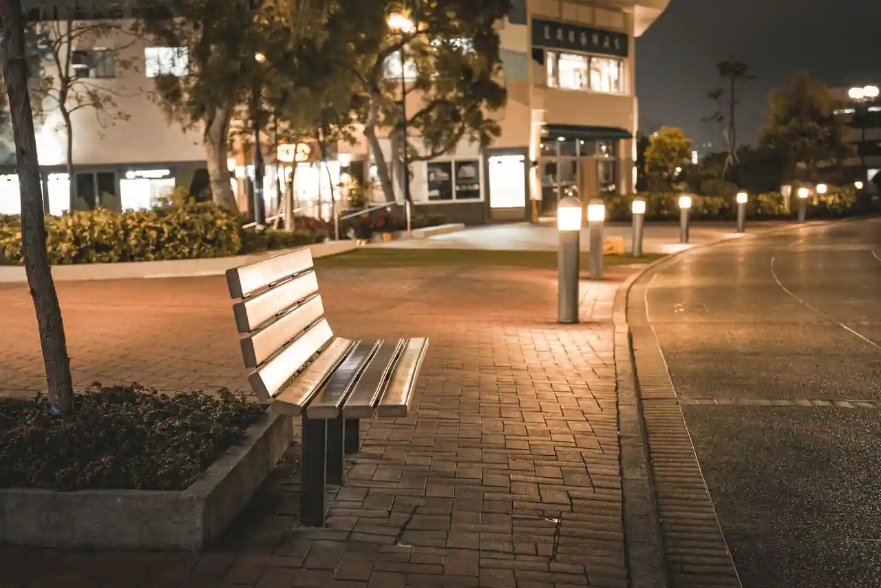 Banc public vide, bornes lumineuses et jardinière le long d’un trottoir pavé de centre-ville, le soir