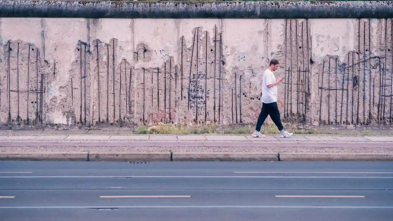 Vestige du mur de Berlin au béton buriné, armatures d’acier à nu, un passant longeant la paroi.