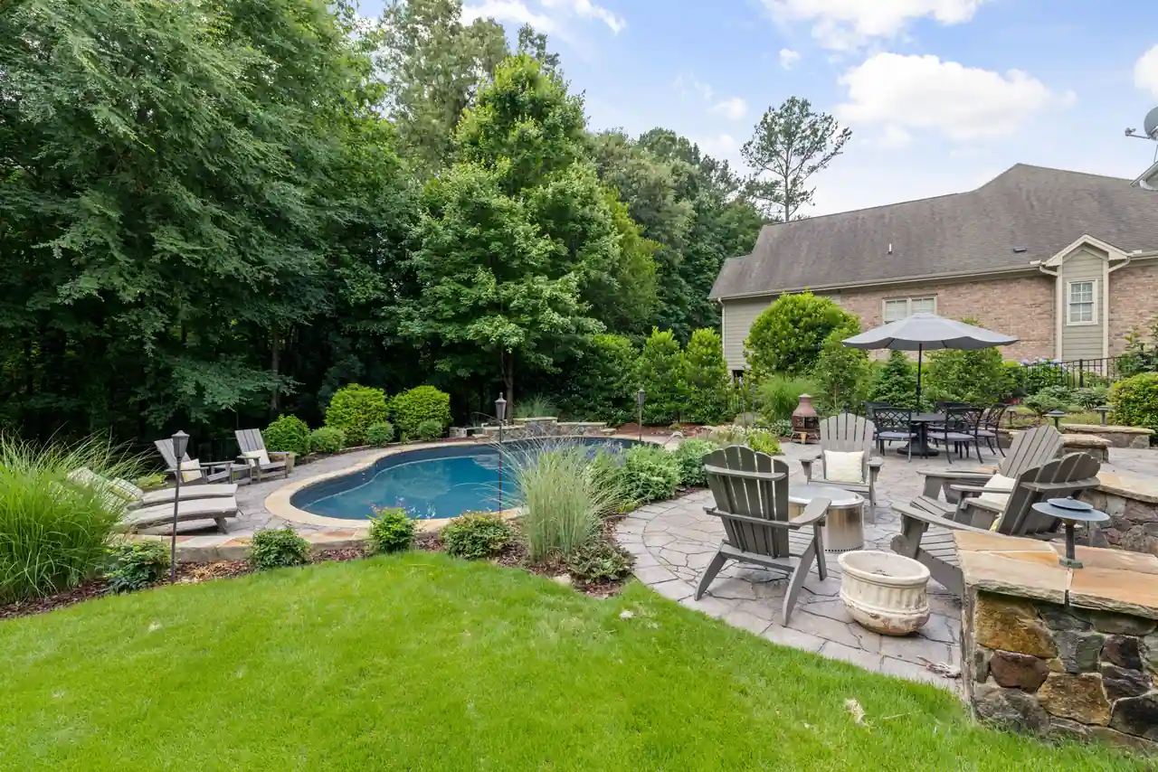 Piscine de forme libre bordée d’une terrasse en pierre, dans un jardin arboré à l’arrière d’une maison