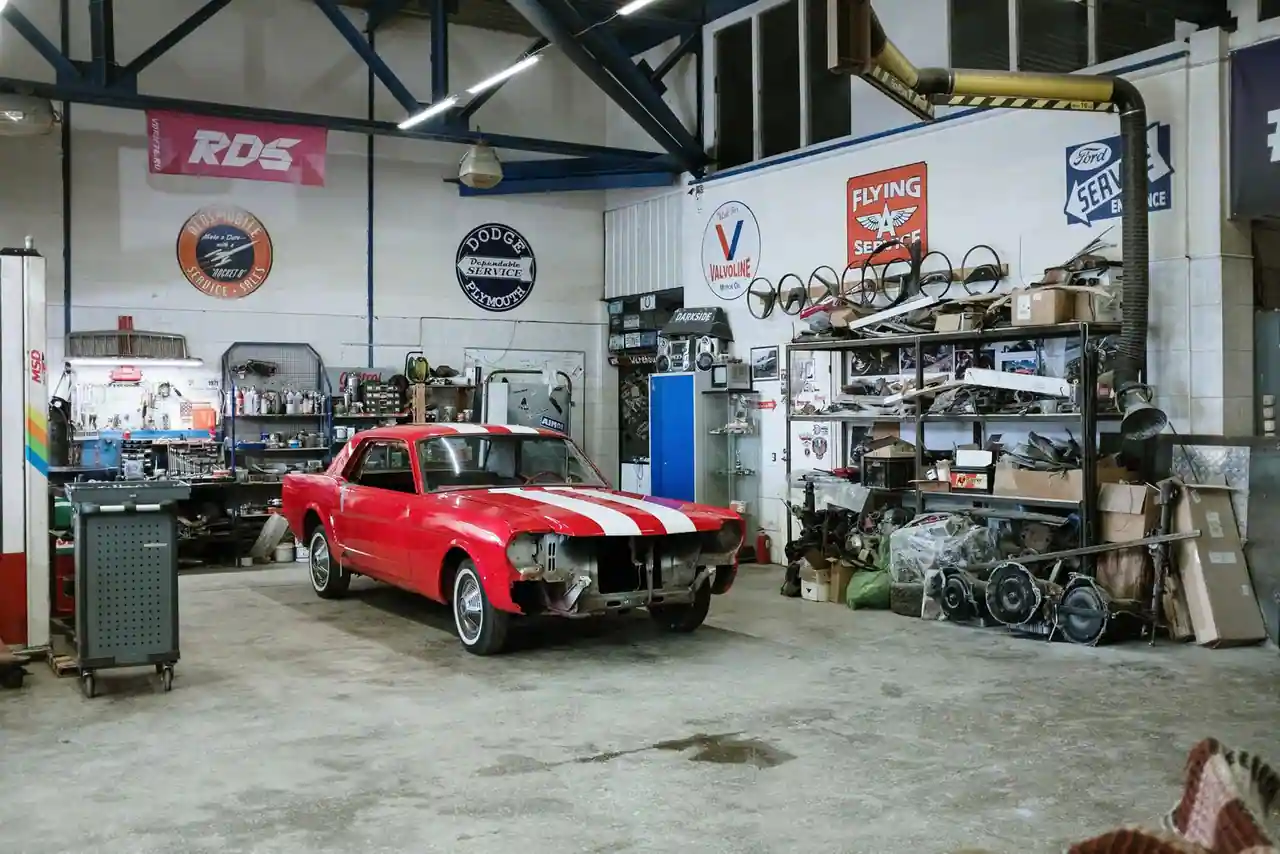 Voiture ancienne rouge à bandes blanches, calandre déposée, dans un atelier garni d'étagères de pièces.