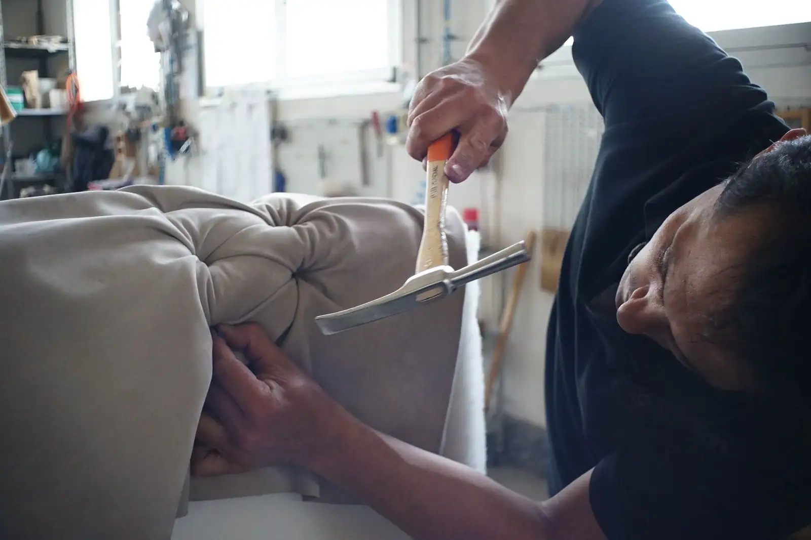 Tapissier au travail sur une assise rembourrée beige, maillet et outil de dégarnissage en main, en atelier.