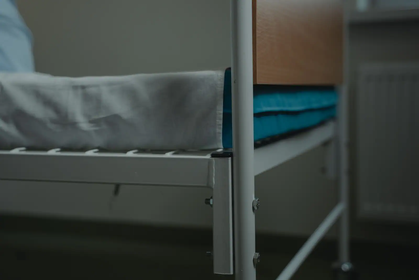 Pied d'un lit d'hôpital à cadre métallique, drap blanc et housse bleue, devant une table de chevet en bois.