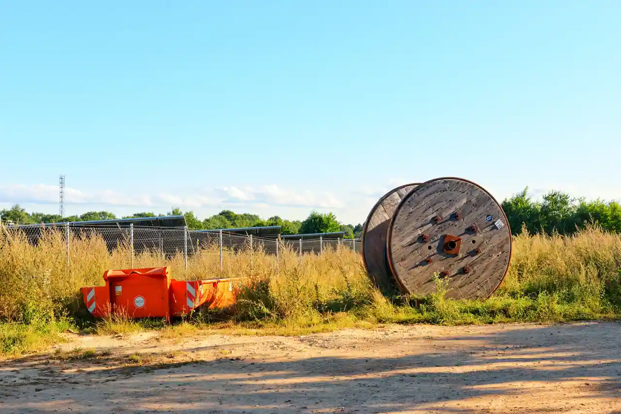 Grand touret de câble en bois posé au sol près d'un bac de chantier orange, en bordure d'un terrain