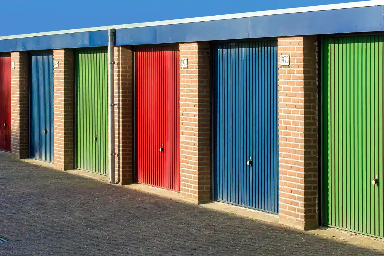 Une rangée de box fermés, portes basculantes colorées et piliers de brique, le long d'une allée.