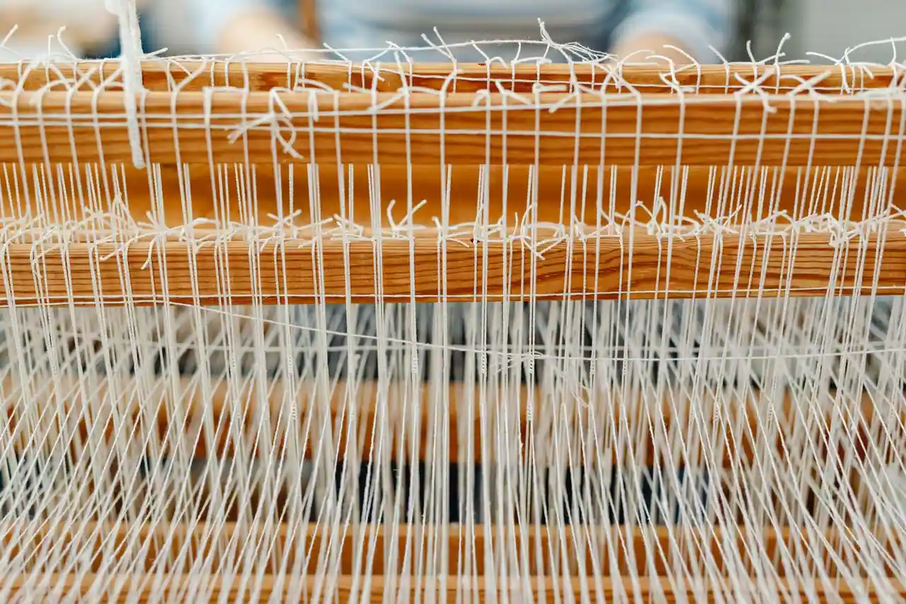 Fils de chaîne blancs tendus sur un métier à tisser en bois, cordelettes de lices nouées sur les barres.