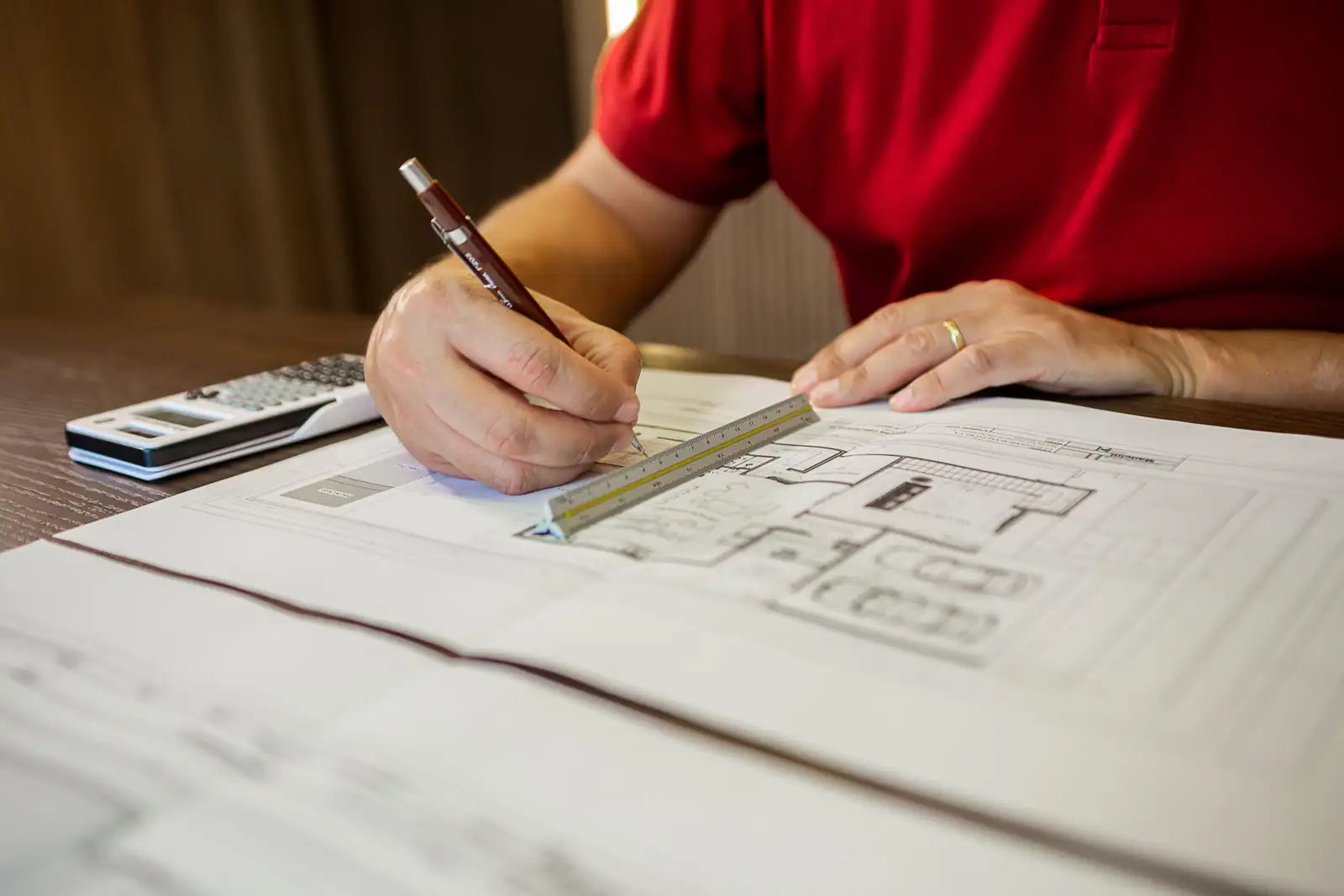 Une personne trace un plan de maison à la règle, avec une calculatrice posée sur la table.